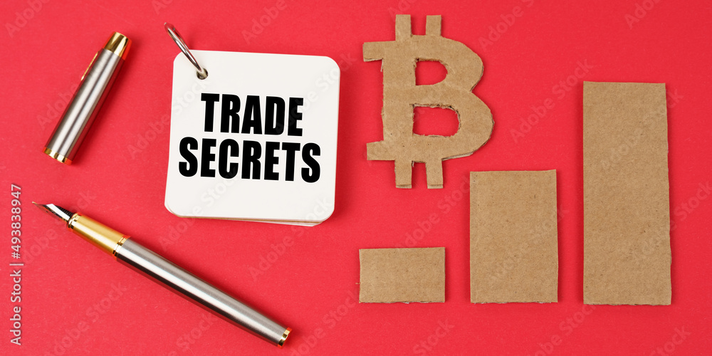 Poster on a red surface lie the bitcoin symbol, a pen and a notepad with the inscription - trade secrets