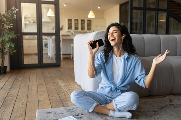 Fototapeta premium Cheery young woman in earphones listening to music, singing and using smartphone as mic, sitting on floor at home