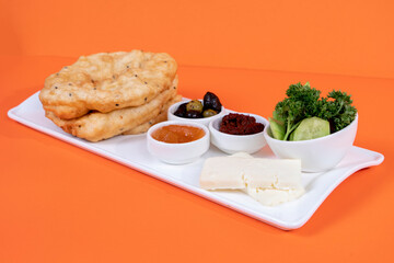 Cropped view of greens platter, tomato paste, olives, citrus jam, cheese slices and bagels on white plate, shot with selective focus on orange background in isolated setting.