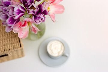 A bouquet of open pink and purple tulips, a wicker basket, a white cup with coffee on a white table.