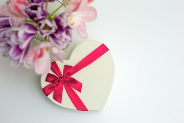 A bouquet of open pink and purple tulips , a heart-shaped gift box, with a pink bow, on a white table .