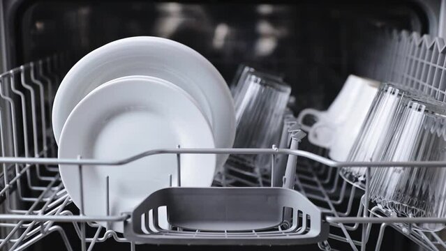 Closeup of woman hands insert and load basket, puts a washed plate to opened automatic stainless built-in dishwasher machine with clean utensils inside in modern home kitchen. Household domestic life