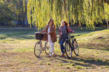 Two young woman with bikes walk and talk and smile in public park