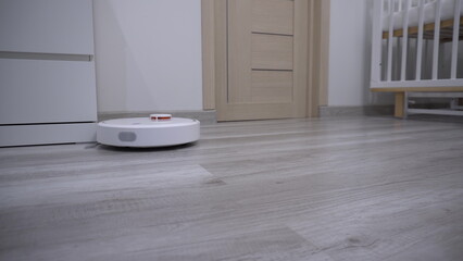 A white robot vacuum cleaner rides on a white parquet floor. The robot with artificial intelligence determines the space and cleans the floor.