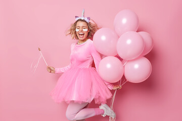 Obraz na płótnie Canvas Positive cheerful young woman exclaims from joy keeps mouth widely opened poses with bunch of inflated balloons dances and has fun moves energetic against pink background. Party time concept