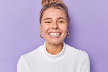 Portrait of cheerful young female model smiles broadly shows perfect teeth wears golden earrings and casual white jumper looks gladfully poses against purple background. Happy emoions concept