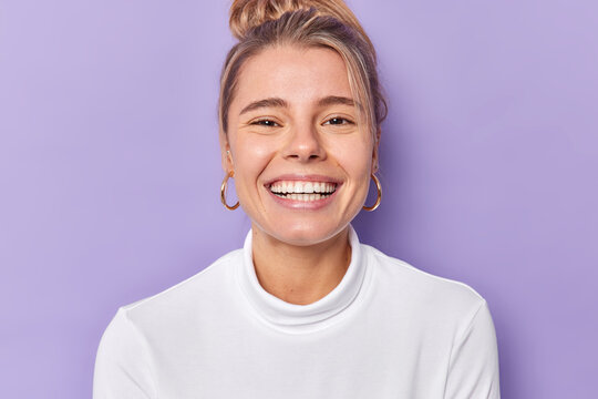 Portrait of cheerful young female model smiles broadly shows perfect teeth wears golden earrings and casual white jumper looks gladfully poses against purple background. Happy emoions concept