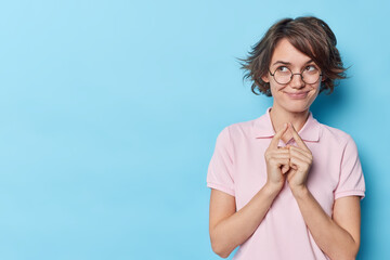 Pretty cunning girl with bob hairstyle has idea makes plan schemes something smiles pleasantly dressed in casual t shirt and round spectacles isolated over blue background blank space for your promo