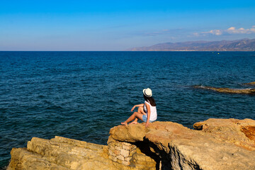 Fototapeta na wymiar girl in a white hat sits on a rocky seashore, a beautiful view of the sea in Crete