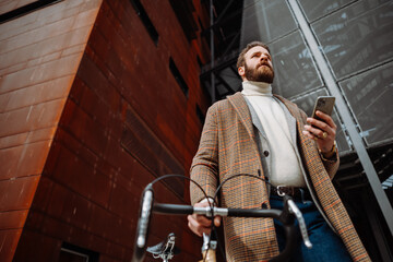 Young man with bicycle holding a smartphone front of building. Creative businessman in a modern business area.
