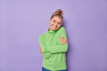 Pleased lovely woman with combed hair embraces herself feels comfortable wears green jumper smiles gently being self egoist poses against purple background. Self love and acceptance concept.