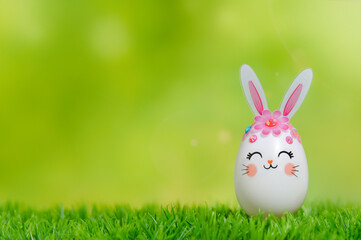 Easter Egg bunny on the grass. Happy Easter composition with place for text. An egg decorated with ears and a muzzle of a rabbit against a background of green spring bokeh. 