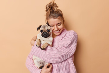 Display of affection. Happy young woman with combed hair embraces pug dog has fun being mom of puppy wears knitted sweater isolated over beige background. Female pet lover with domestic animal