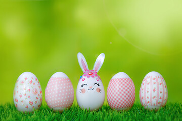 A row of Easter eggs on the grass. Bunny egg and pink-decorated eggs on a green bokeh background. Happy Easter composition with copy space.