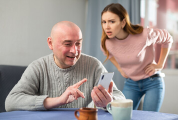Man sitting at table at home and using smartphone. His jealous wife standing behind.