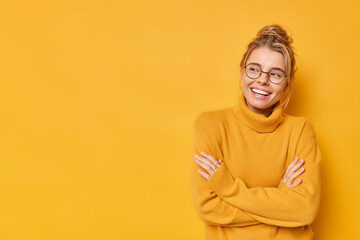 Studio shot of positive young woman with fair combed hair keeps arms folded feels satisfied looks at something wears sweater and spectacles isolated over yellow backgroud blank space for your text