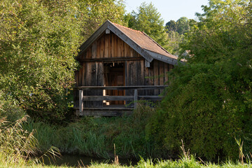 Small brown wooden barn between green trees on the bank of a small river in a cozy Bavarian village on a sunny day