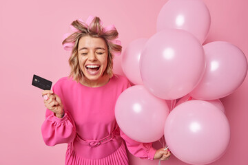 Fototapeta na wymiar Glad young woman with leaked makeup holds credit bank card and bunch of helium balloons glad to celebrate birthday and spends money for party wears dress makes hairstyle isolated over pink background