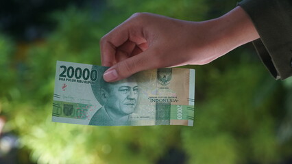 The Indonesian currency, the 20,000 rupiah note, is held in a woman's hand