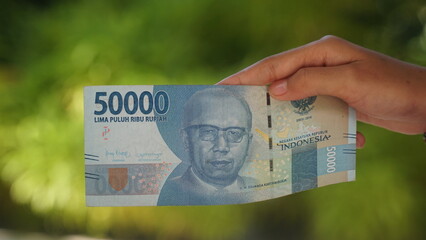 The Indonesian currency, the 50,000 rupiah note, is held in a woman's hand