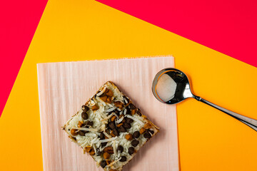 Top down view of layered pastry garnished with coconut pills, chocolate chip and pecan peaces placed on baking sheet on bright orange and pink background. Shiny spoon placed next to it. 