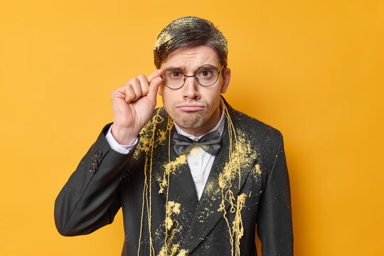 Horizontal Shot Of Attentive Serious Man Keeps Hand On Rim Of Spectacles Dressed In Festive Suit Smeared With Serpantine Spray Has Bad Mood Isolated Over Yellow Background Upset To Get Older