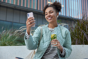 Outdoor shot of happy teenage girl with two curly buns takes selfie via smartphone drinks detox...