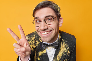 Pleased cheerful adult man smiles gladfully being in good mood wears round spectacles and festive black formal suit enjoys celebration isolated over yellow studio wall. Hey stay always positive