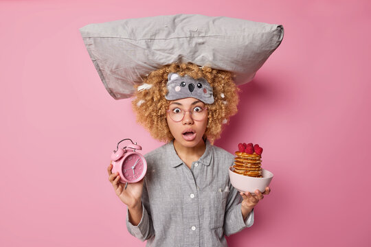 Scared curly woman poses with pillow on head reacts shocked on embarrassing news in morning wears sleepmask and slumber suit holds alarm clock and delicious pancakes awakes early poses indoor