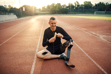 A happy runner with artificial leg listening music, and peeling banana while relaxing at stadium.