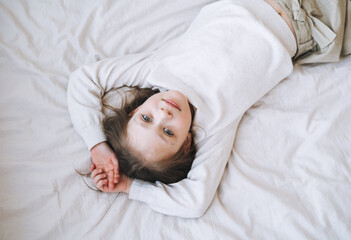 Obraz premium Cuty funny little girl with long hair in white home dress having fun lies on bed at the home, view from top