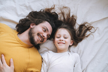 Young happy family with bearded father and daughter on bed in cozy home, view from top