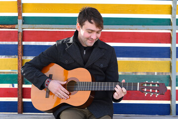 Smiling young man playing guitar and sitting against colorful wall