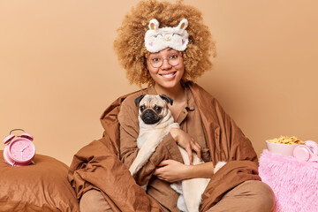Positive smiling curly haired woman wrapped in blanket carries pug dog on hands stays in bed going to have sleep enjoys cozy domestic atmosphere poses against beige background. Family time concept