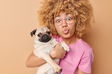 Lovely curly haired young woman keeps lips folded embraces pug dog has new family member wears transparent eyeglasses and t shirt isolated over beige background. People and domestic animals.
