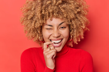 Positive curly haired young woman bites finger winks eye and smiles broadly shows white teeth being in good mood wears golden earrings casual turtleneck isolated over red background. Happy emotions