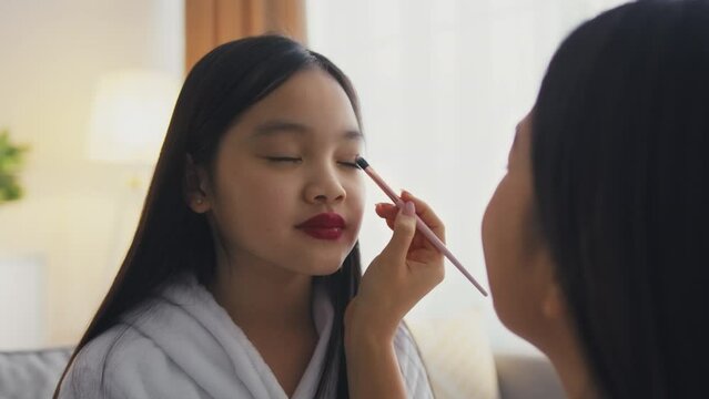 Loving mother making professional make up for her little asian daughter, playing beauty salon at home, slow motion