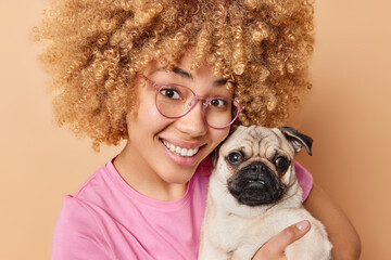 Pretty curly millennial girl with glad expression poses with favorite pug dog spends free time with lovely puppy smiles broadly going to have walk wears spectacles casual clothes poses indoor