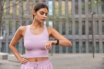 Fototapeta premium Fit healthy brunette young woman dressed in sportswear checks fitness results via smartwatch keeps hand on waist goes in for sport outdoors during summer time poses against modern city building