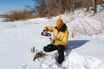 A young man in warm clothes drinks tea in nature. The hand pours a hot drink from a thermos into a mug. Camping camping in the woods, freezing day, survival.