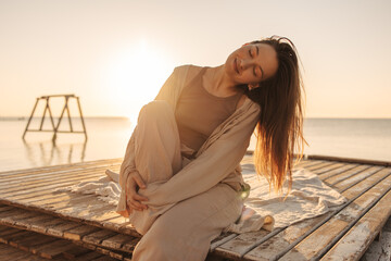 Relaxed young fair-skinned girl sits on pier with closed eyes on ocean shore. Model has leg pressed...
