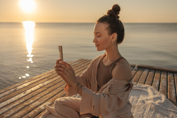 Gentle image of young caucasian girl sitting on wooden pier with gadget in hands on seashore. Brown-haired teenager with bun of hair, dressed light brown spring suit. Concept of solitude with nature.
