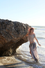 Full length portrait of  red haired woman wearing  torn shipwrecked clothing. Standing  pose with gestural hands at  rocky ocean beach landscape background.
