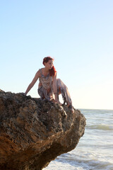 Fototapeta na wymiar Full length portrait of red haired woman wearing torn shipwrecked clothing. Standing pose with gestural hands at rocky ocean beach landscape background. 