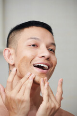 Happly young man applying red clay mask on chin to deep clean pores