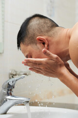 Young man rinsing face with tap water to wash off cleanser in bathroom in the morning