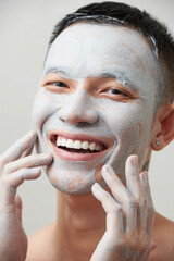 Portrait of joyful young man applying pore tightening clay mask on face