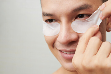 Close-up image of smiling young man applying patches to brighten and reduce dark circles under eyes