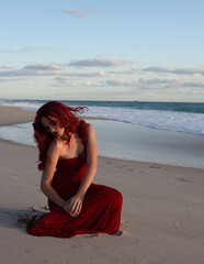 Fototapeta na wymiar Full length portrait of redhead woman wearing elegant gown. Standing pose with gestural hands at sunset ocean beach landscape background.