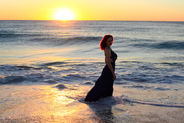 Full length portrait of  redhead woman wearing elegant gown. Standing  pose with gestural hands at sunset ocean beach landscape background.